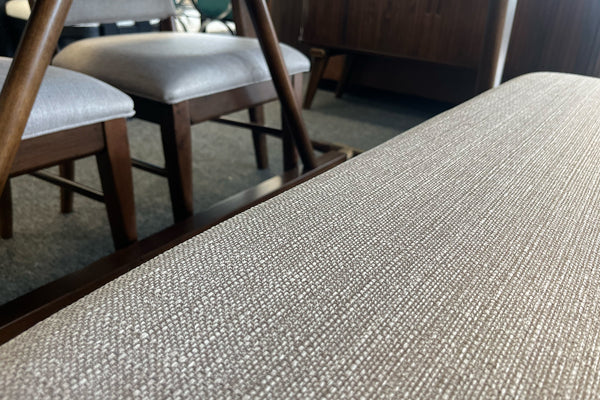Primer plano de un banco tapizado en tela beige texturizada con estructura de madera oscura. Al fondo se observan sillas de comedor a juego en una sala de exhibición, destacando los materiales y acabados del conjunto.