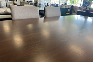 Primer plano de una mesa de comedor rectangular con acabado en madera oscura y superficie lisa. Al fondo se observan dos sillas tapizadas en tela beige dentro de una sala de exhibición de muebles con iluminación natural.