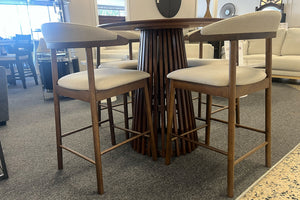 Juego de comedor tipo pub con mesa redonda de madera oscura y base pedestal de listones verticales, acompañado de cuatro sillas altas tapizadas en beige. Exhibido en una sala de muebles con diseño moderno y acabados cálidos.
