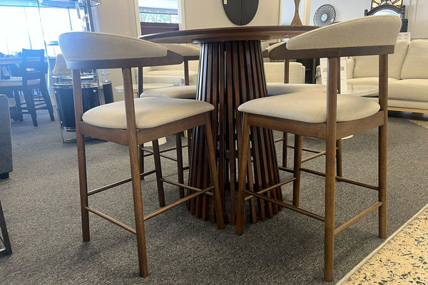 Juego de comedor tipo pub con mesa redonda de madera oscura y base pedestal de listones verticales, acompañado de cuatro sillas altas tapizadas en beige. Exhibido en una sala de muebles con diseño moderno y acabados cálidos.