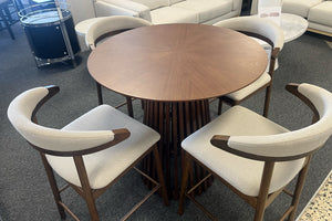 Juego de comedor tipo pub con mesa redonda de madera oscura y base pedestal de listones verticales, rodeada por cuatro sillas altas con estructura de madera y respaldo curvo tapizado en tela beige. Exhibido en una sala de muebles moderna.