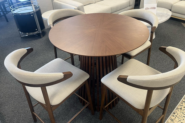 Juego de comedor tipo pub con mesa redonda de madera oscura y base pedestal de listones verticales, rodeada por cuatro sillas altas con estructura de madera y respaldo curvo tapizado en tela beige. Exhibido en una sala de muebles moderna.