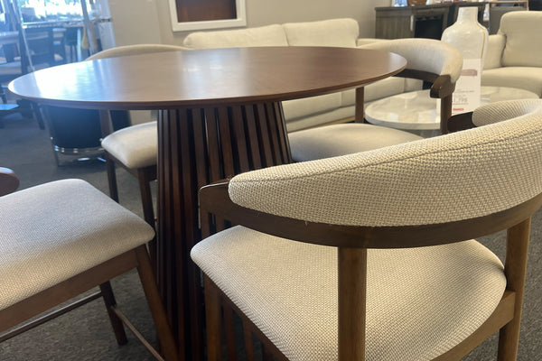 Juego de comedor tipo pub con mesa redonda de madera oscura y base pedestal de listones verticales. Incluye cuatro sillas altas con estructura de madera y respaldo curvo tapizado en tela beige, exhibido en una sala de muebles contemporánea.