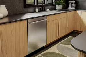 Una escena de cocina moderna que muestra un lavaplatos bajo encimera de acero inoxidable instalado en gabinetes de madera clara junto a una encimera oscura.