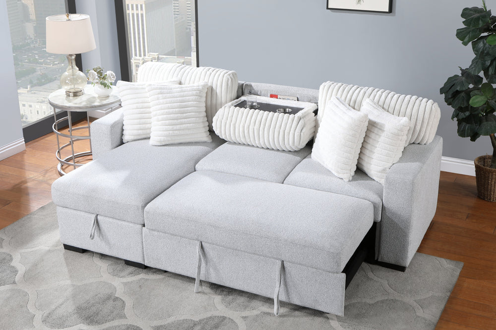 Sofá chaise de color gris claro con cojines blancos en una sala con objetos decorativos y ventanas en el fondo. 