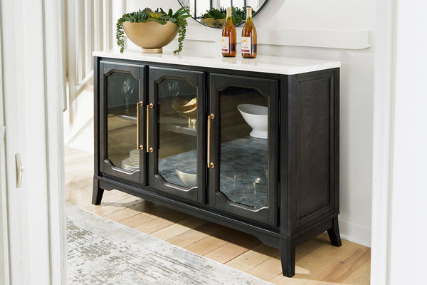 Un servidor de madera negra con tres puertas de frente acristalado, tiradores dorados y encimera de mármol blanco, situado en un vestíbulo luminoso y decorado con objetos ornamentales sobre la superficie.