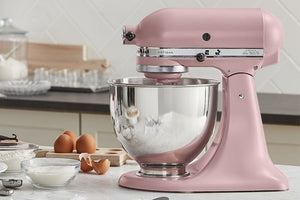 Una batidora KitchenAid Artisan rosa con un bol de acero inoxidable lleno de harina blanca, colocada sobre una encimera de cocina con huevos, boles con ingredientes y un fondo de azulejos.