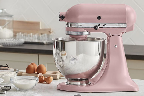 Una batidora KitchenAid Artisan rosa con un bol de acero inoxidable lleno de harina blanca, colocada sobre una encimera de cocina con huevos, boles con ingredientes y un fondo de azulejos.