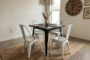 Mesa de comedor cuadrada con superficie de madera oscura y estructura metálica negra, acompañada por cuatro sillas blancas de estilo industrial. El conjunto está sobre una alfombra tejida en un comedor moderno con lámparas colgantes y reloj decorativo de pared.