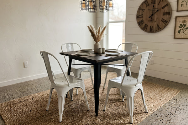 Mesa de comedor cuadrada con superficie de madera oscura y estructura metálica negra, acompañada por cuatro sillas blancas de estilo industrial. El conjunto está sobre una alfombra tejida en un comedor moderno con lámparas colgantes y reloj decorativo de pared.