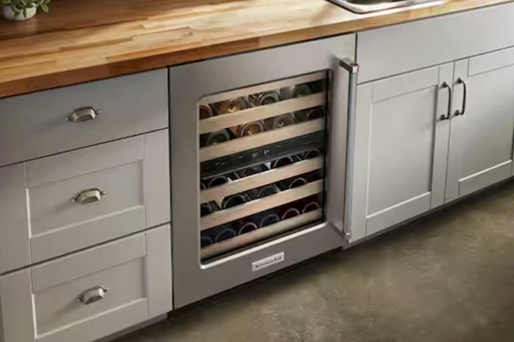 Una nevera para vinos KitchenAid bajo encimera con exterior de acero inoxidable, tirador vertical y puerta de cristal que deja ver listones de madera horizontales y negros en el interior.