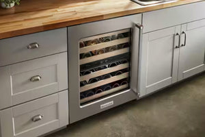 Una nevera para vinos de acero inoxidable bajo la encimera, con la puerta abierta, que deja ver botellas de vino en estantes de madera dentro de un armario de cocina, junto a cajones blancos y una encimera de madera.