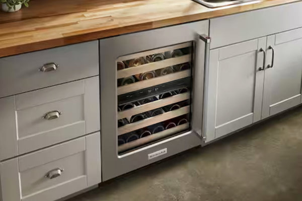 Una nevera para vinos de acero inoxidable bajo la encimera, con la puerta abierta, que deja ver botellas de vino en estantes de madera dentro de un armario de cocina, junto a cajones blancos y una encimera de madera.