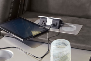 Primer plano de consola integrada en sofá reclinable tapizado en cuero sintético gris oscuro, con enchufe eléctrico, puerto USB, base de carga inalámbrica y tableta conectada mediante cable de carga.
