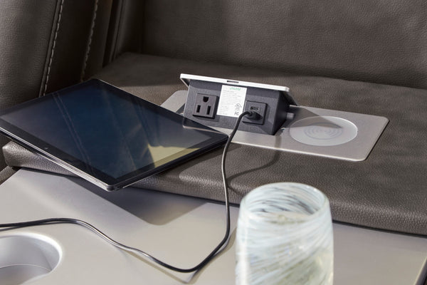Primer plano de consola integrada en sofá reclinable tapizado en cuero sintético gris oscuro, con enchufe eléctrico, puerto USB, base de carga inalámbrica y tableta conectada mediante cable de carga.
