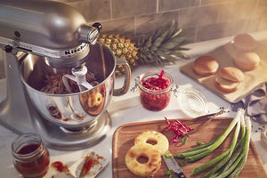 Una batidora KitchenAid reposa sobre la encimera de la cocina, rodeada de ingredientes y alimentos preparados, como piña, cebolletas, aros de pan y panecillos para hamburguesas.