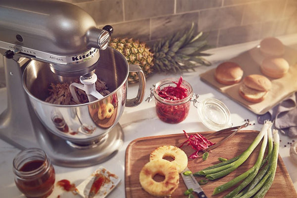 Una batidora KitchenAid reposa sobre la encimera de la cocina, rodeada de ingredientes y alimentos preparados, como piña, cebolletas, aros de pan y panecillos para hamburguesas.