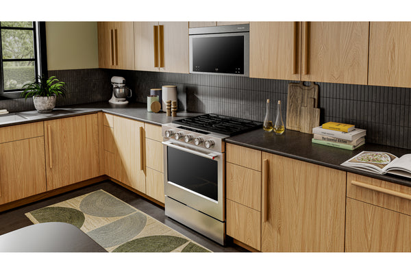 Un interior de cocina moderno con gabinetes de madera clara, encimeras oscuras, una estufa independiente de acero inoxidable, microondas negro empotrado, protector contra salpicaduras de azulejos oscuros y artículos de cocina decorativos que incluyen una batidora de pie, tablas de cortar, libros y una planta.