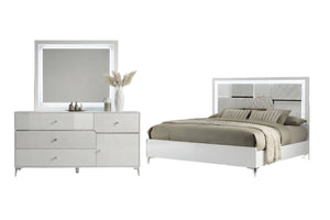 Un moderno conjunto de muebles de dormitorio que incluye un gavetero blanco de seis gavetas con espejo rectangular incorporado y una cama blanca a juego con cabecero geométrico, todo ello decorado con ropa de cama beige sobre un fondo blanco liso.