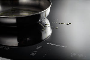 Un primer plano de un tope de inducción KitchenAid negra con una sartén de acero inoxidable encima, que muestra algunas gotas de aceite en la superficie y la marca impresa en la placa.