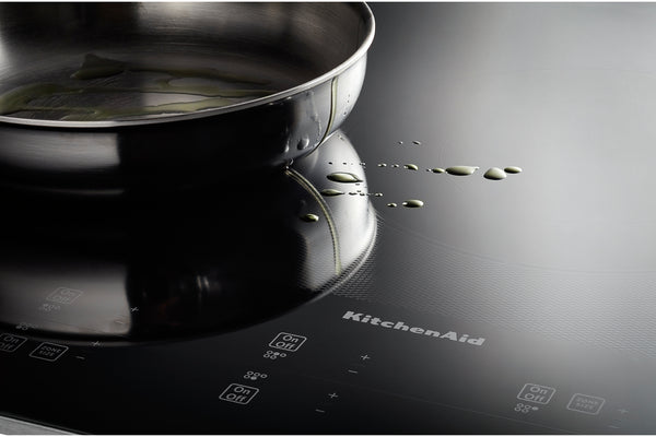 Un primer plano de un tope de inducción KitchenAid negra con una sartén de acero inoxidable encima, que muestra algunas gotas de aceite en la superficie y la marca impresa en la placa.