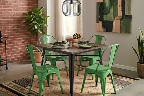 Mesa de comedor cuadrada con cubierta de madera oscura y patas metálicas negras, acompañada por cuatro sillas verdes de estilo industrial. El conjunto está sobre una alfombra decorativa en un comedor moderno con pared de ladrillo, plantas y lámpara colgante.
