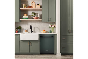 Una escena de cocina moderna que presenta un lavavajillas integrado de color verde oscuro debajo de un fregadero blanco de estilo rústico, con gabinetes a juego de color verde oscuro, estantes flotantes de madera clara y un protector contra salpicaduras de azulejos de color gris claro.