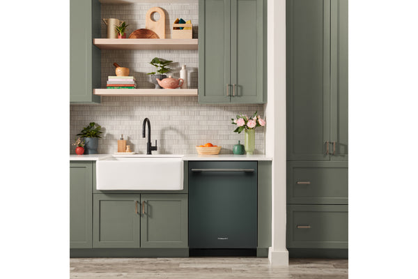 Una escena de cocina moderna que presenta un lavavajillas integrado de color verde oscuro debajo de un fregadero blanco de estilo rústico, con gabinetes a juego de color verde oscuro, estantes flotantes de madera clara y un protector contra salpicaduras de azulejos de color gris claro.