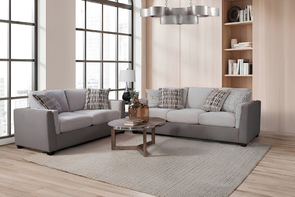 Conjunto de sala moderna con un sofás y un loveseat de tela en tonos gris y beige, cada uno con cojines acolchados y almohadas decorativas con patrón neutro. Los muebles están colocados en ángulo sobre una alfombra clara, alrededor de una mesa de centro redonda con libros y objetos decorativos. El espacio incluye una mesa auxiliar con lámpara, estantería con libros y una gran ventana que aporta luz natural, mostrando un ambiente contemporáneo y luminoso.