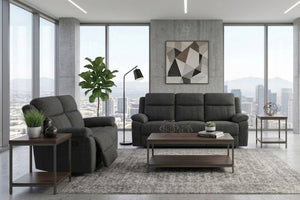 Un moderno salón con un sofá gris oscuro y un loveseat reclinable a juego sobre una alfombra estampada, con una mesa de centro de madera al frente. La habitación cuenta con grandes ventanales con vistas al horizonte de la ciudad, una pared de hormigón con un mural geométrico y una planta en maceta.