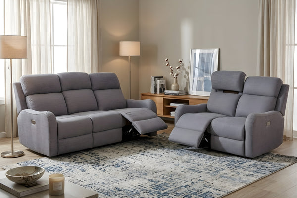 Sala contemporánea con sofá y loveseat reclinables en color gris claro, ambos con reposapiés extendidos. El loveseat incluye cabeceras ajustables y el espacio está decorado con lámparas, alfombra estampada y mesa auxiliar.