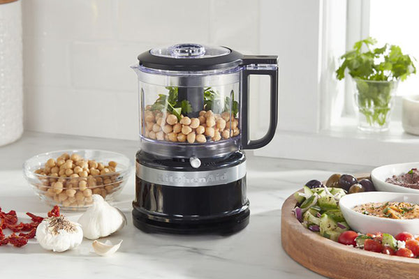 Un procesador de alimentos KitchenAid negro lleno de garbanzos y verduras, colocado sobre una encimera de mármol blanco en una cocina luminosa con cuencos de ingredientes cerca.