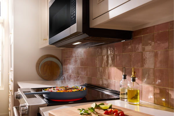 Una escena de cocina moderna con una campana extractora de acero inoxidable y color negro montada bajo gabinetes blancos, un salpicadero de azulejos en tonos cálidos de marrón rosado, una sartén con comida cocinándose en la estufa y botellas de aceite sobre la encimera.