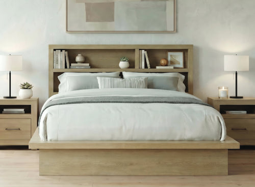 Cama con plataforma de roble claro y cabecero con librería integrada, vestida con ropa de cama blanca y cojines grises, flanqueada por mesitas de noche a juego con lámparas en un dormitorio luminoso.