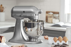 Una batidora de pie KitchenAid Artisan Series en color plata cepillada, mostrada sobre una encimera de cocina con un bol de acero inoxidable, rodeada de ingredientes para hornear como huevos, pan y chocolate.