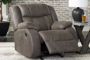 Una butaca reclinable mullido y tapizado en una cálida tela marrón topo, que se muestra reclinado con el reposapiés extendido. El sillón está ubicado en una habitación decorada con una pared de ladrillo visto, una mesa auxiliar con una lámpara y una alfombra estampada.