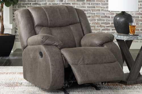 Una butaca reclinable mullido y tapizado en una cálida tela marrón topo, que se muestra reclinado con el reposapiés extendido. El sillón está ubicado en una habitación decorada con una pared de ladrillo visto, una mesa auxiliar con una lámpara y una alfombra estampada.