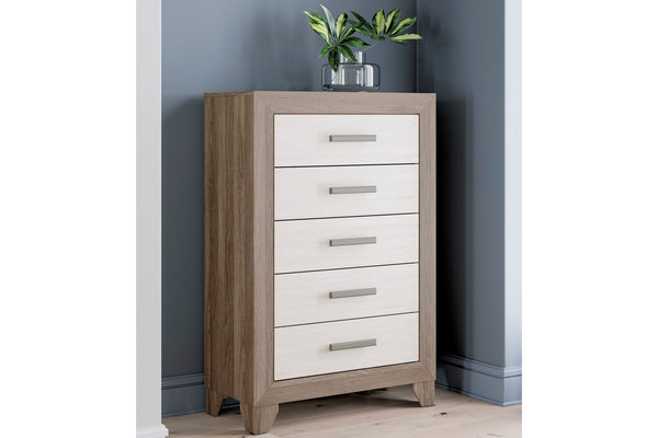 Un chest de 5 cajones con estructura de veta de madera café clara y frentes de cajón blancos, ambientado en un dormitorio con una planta en la parte superior.