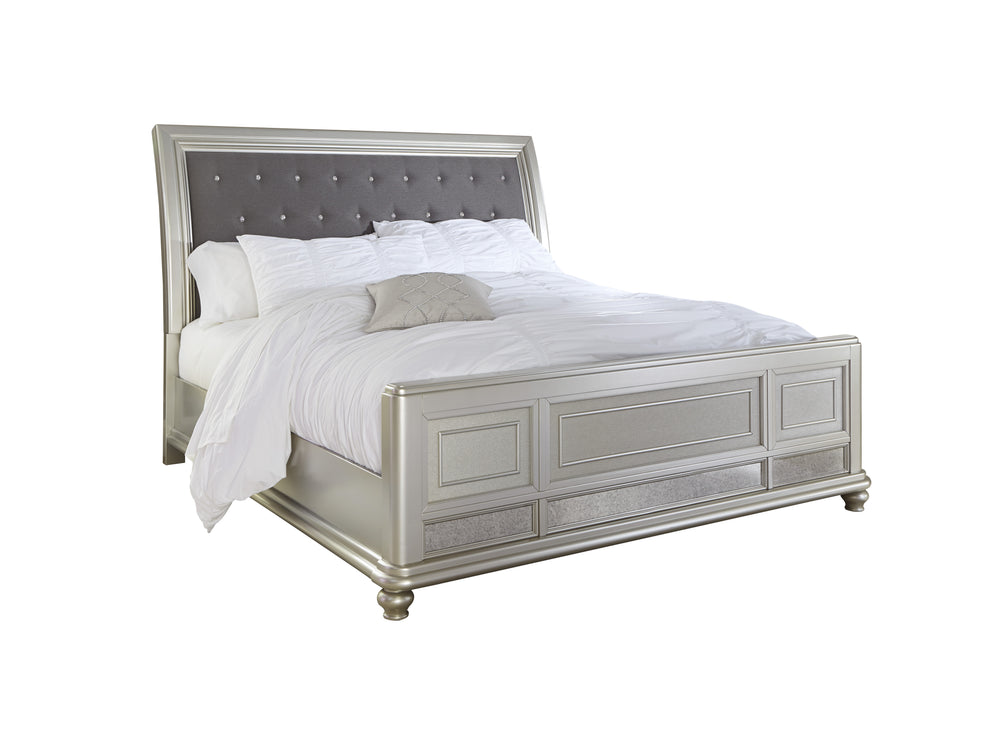 Una estructura de cama con acabado plateado, cabecero gris capitoné y ropa de cama blanca, mostrada sobre un fondo blanco liso.