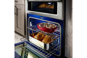 Horno doble empotrado con la puerta del horno inferior abierta, mostrando carnes asadas y una cazuela roja sobre las rejillas. El interior del horno es azul y el exterior tiene un panel frontal de acero inoxidable oscuro.