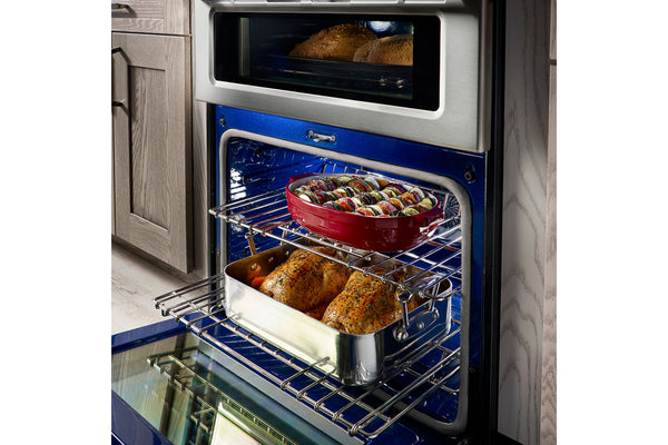 Horno doble empotrado con la puerta del horno inferior abierta, mostrando carnes asadas y una cazuela roja sobre las rejillas. El interior del horno es azul y el exterior tiene un panel frontal de acero inoxidable oscuro.