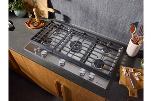 Un tope de gas empotrado con cinco quemadores y mandos plateados, instalada sobre una encimera oscura en una cocina moderna con armarios de madera y un salpicadero de azulejos grises.