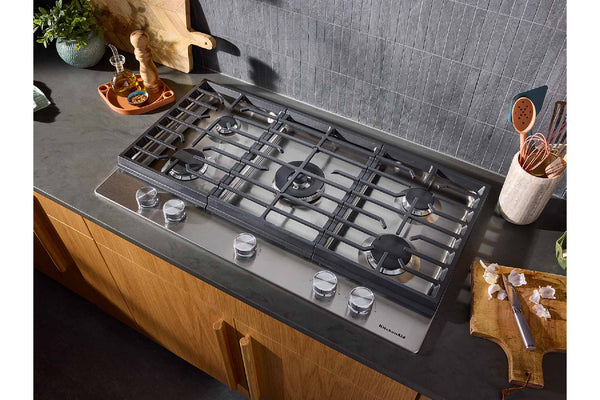 Un tope de gas empotrado con cinco quemadores y mandos plateados, instalada sobre una encimera oscura en una cocina moderna con armarios de madera y un salpicadero de azulejos grises.