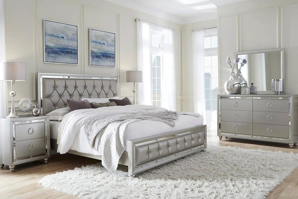 Juego de dormitorio moderno en acabado gris claro con detalles brillantes, presentado sobre fondo blanco. Incluye cama matrimonial con cabecera y pie tapizados en diseño capitoné, gavetero con seis gavetas y tiradores circulares metálicos, espejo rectangular a juego y una mesa de noche de dos gavetas. Diseño contemporáneo con líneas rectas y acabados tipo espejo en los bordes.