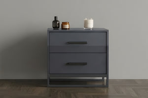 Mesa de noche con dos gavetas de color gris oscuro con manijas negras sobre un fondo neutral de sala. 