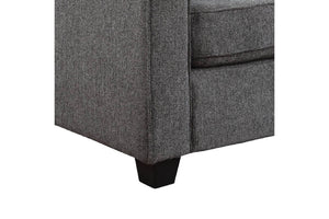 Primer plano de la esquina de un sillón o sofá tapizado en gris con patas de madera oscura, sobre un fondo blanco liso.