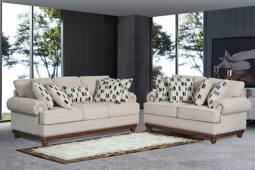 Juego de sala clásico con sofá y loveseat tapizados en tela beige clara, cojines decorativos estampados en blanco, negro y gris, apoyabrazos enrollados con detalles de tachuelas metálicas y base de madera oscura, ubicado en una sala moderna con grandes ventanas y alfombra decorativa.