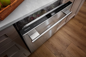 Un lavavajillas empotrado de acero inoxidable con panel de control negro y tirador horizontal largo, instalado bajo una encimera blanca en una cocina con suelo de madera.