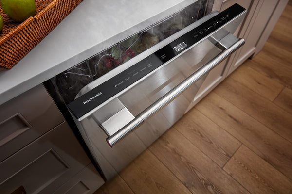Un lavavajillas empotrado de acero inoxidable con panel de control negro y tirador horizontal largo, instalado bajo una encimera blanca en una cocina con suelo de madera.