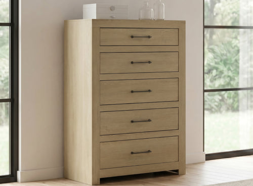Chest de madera clara con 5 gavetas y agarraderas de barra de metal oscuro, ubicada en la ambientación de un dormitorio entre dos ventanas.